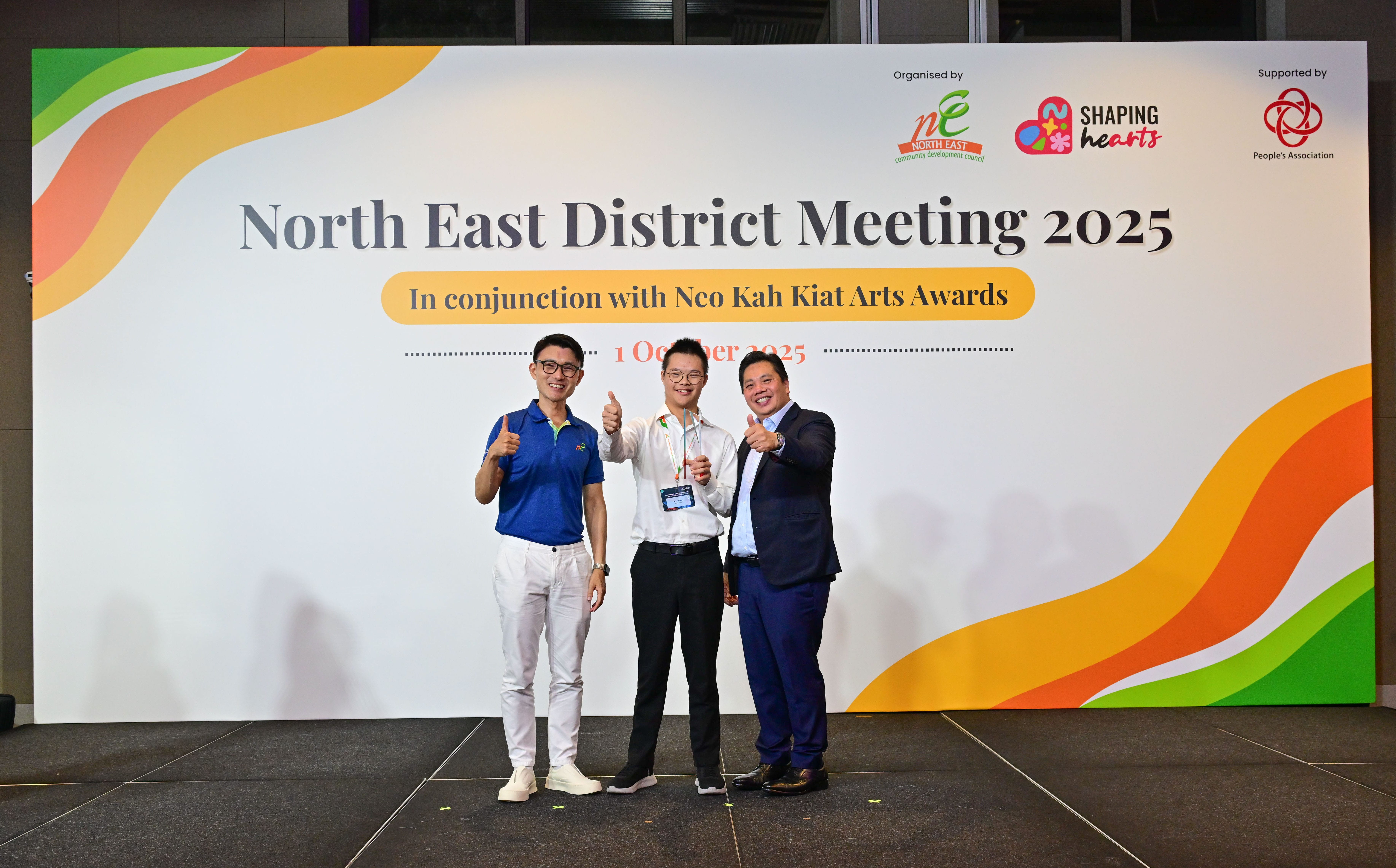 Award recipient standing on stage with the presenter, holding a acrylic plaque and posing for a group photo during the award ceremony, with a backdrop reading ‘North East District Meeting 2025 In conjunction with Neo Kah Kiat Arts Awards'